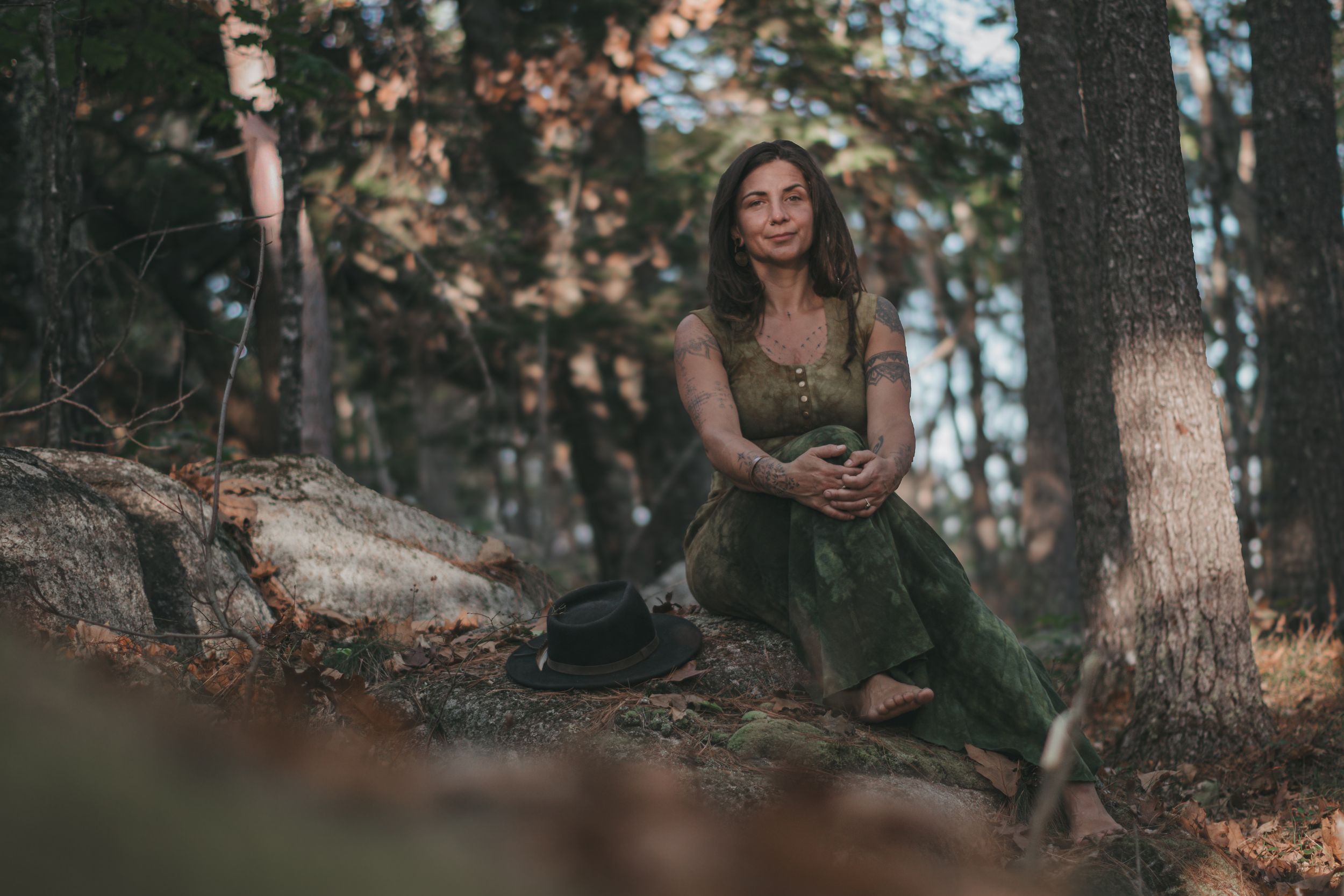 A woman in a green dress sits peacefully on rocks in a forest, with a black hat beside her.