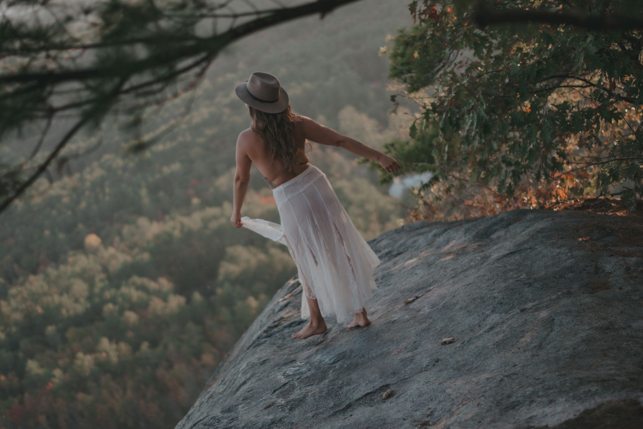 Woman in white dress and hat standing on a rocky cliff edge overlooking a lush forest valley.