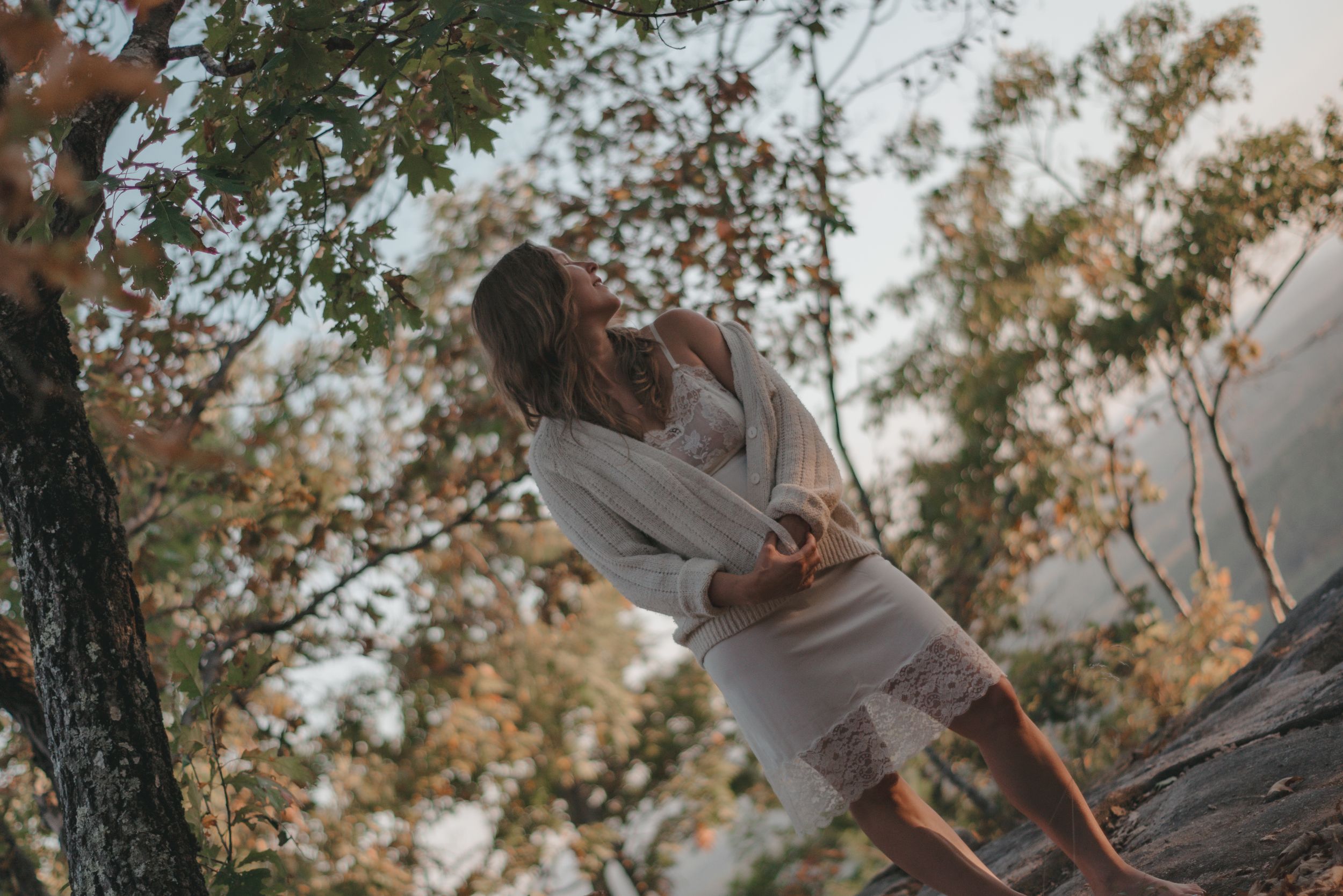 A woman in a white lace dress leans against a tree trunk surrounded by autumn foliage in a serene outdoor setting.
