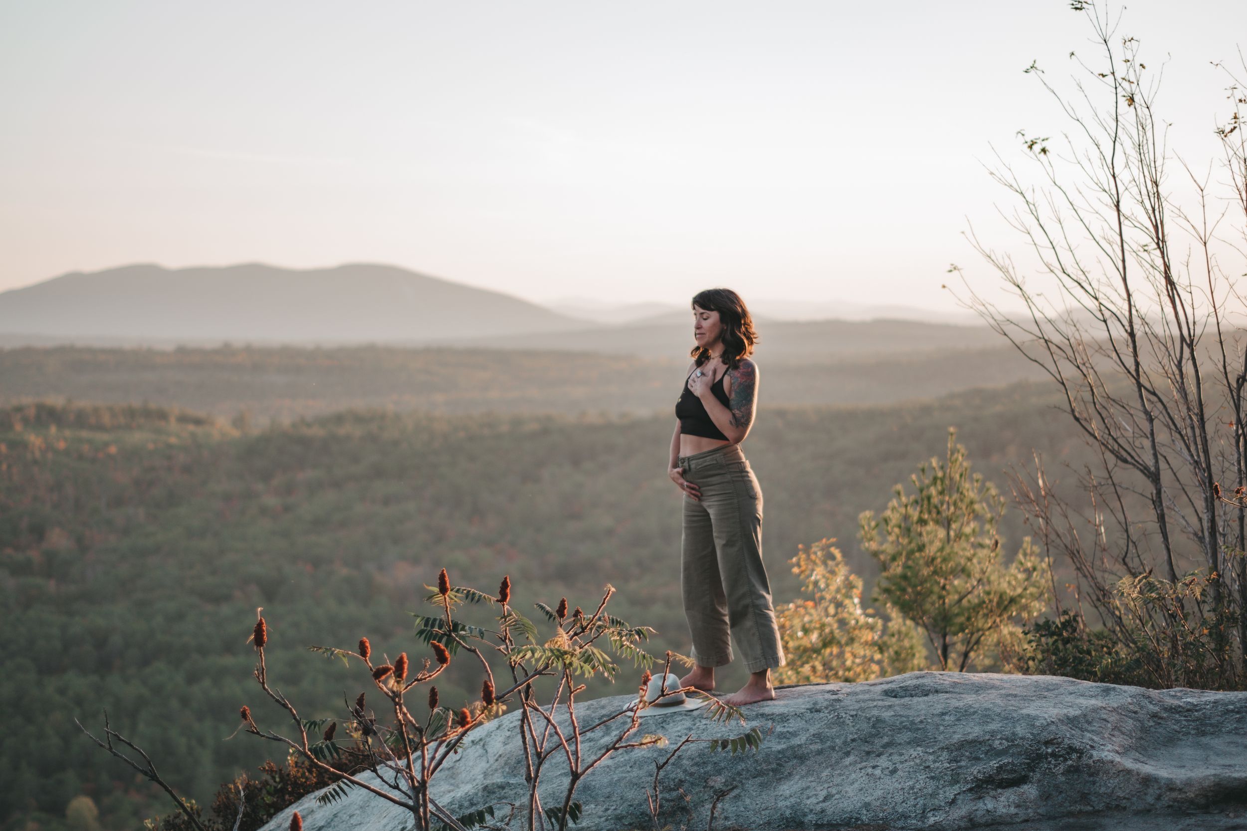 Pregnant woman standing on a rocky overlook with a scenic mountain valley and forest landscape at sunset.