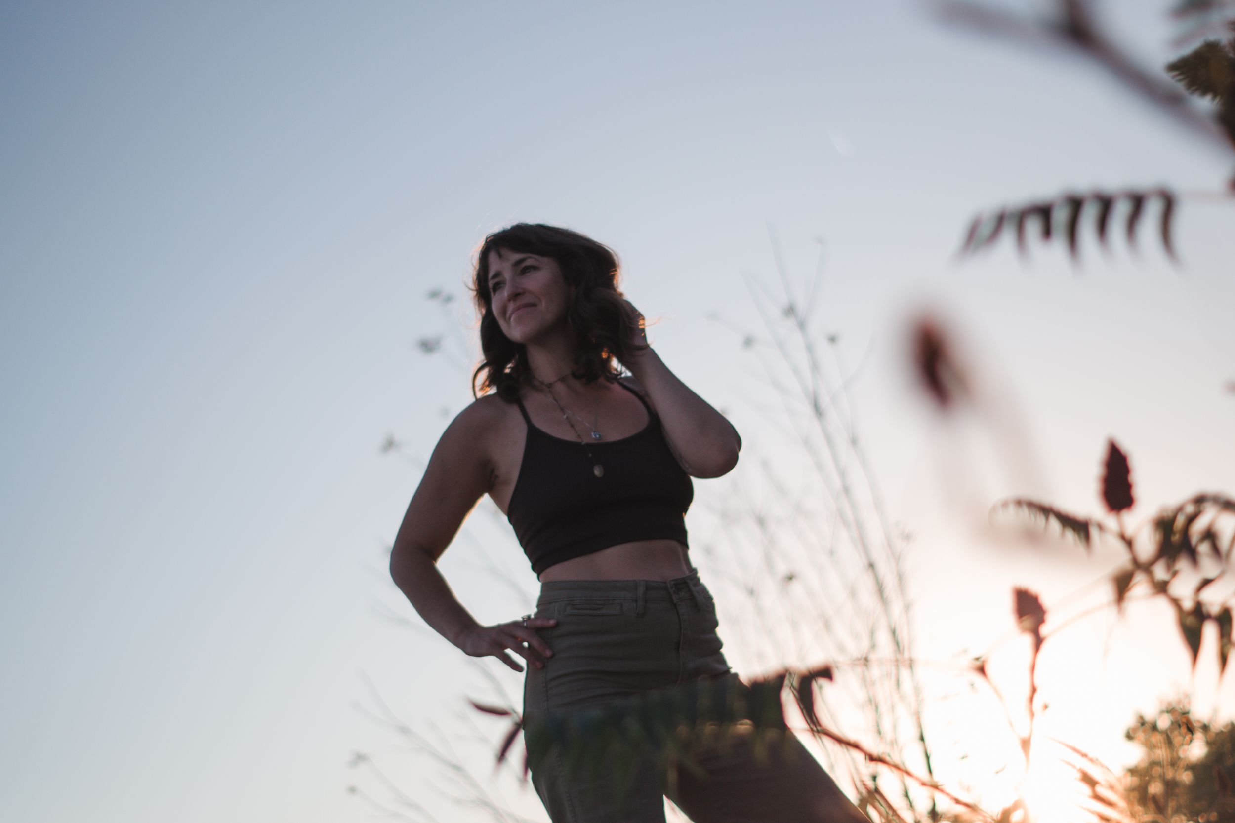 Young woman in black crop top and green pants posing outdoors at sunset with plants in foreground.
