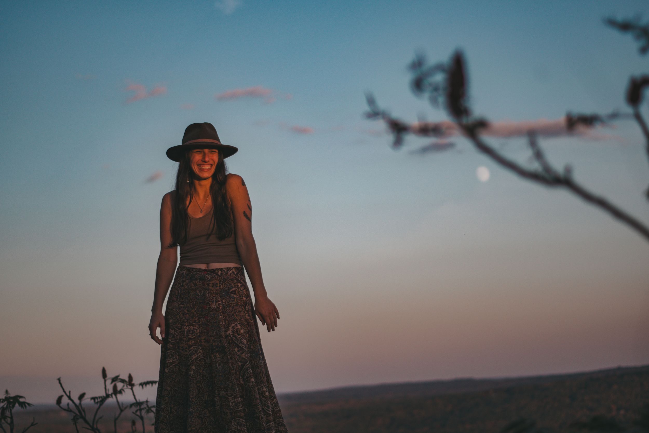 Woman in wide-brim hat and floral skirt stands outdoors at dusk with moon visible in twilight sky.