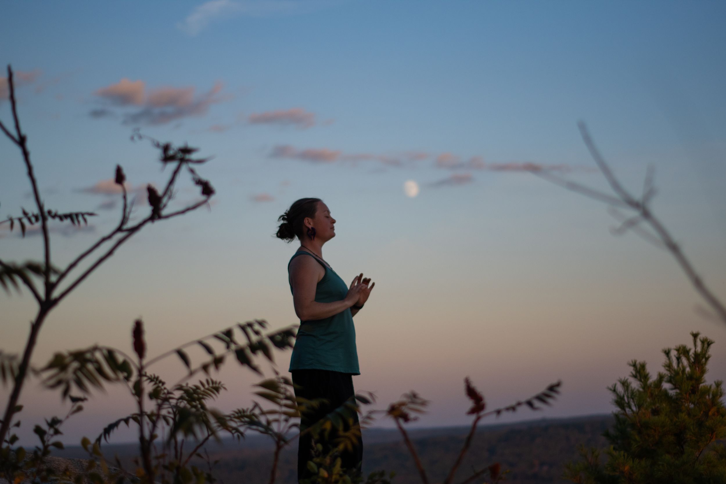 Woman practicing yoga meditation outdoors at dusk with moon visible in a colorful twilight sky.