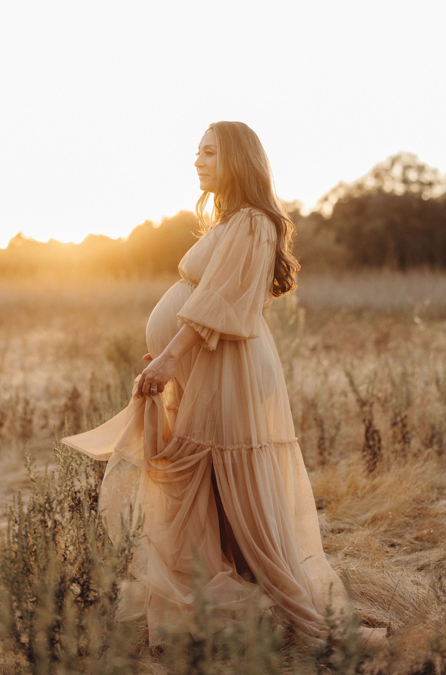 Beautiful maternity photoshoot in a golden field at sunset with flowing cream dress catching the light.