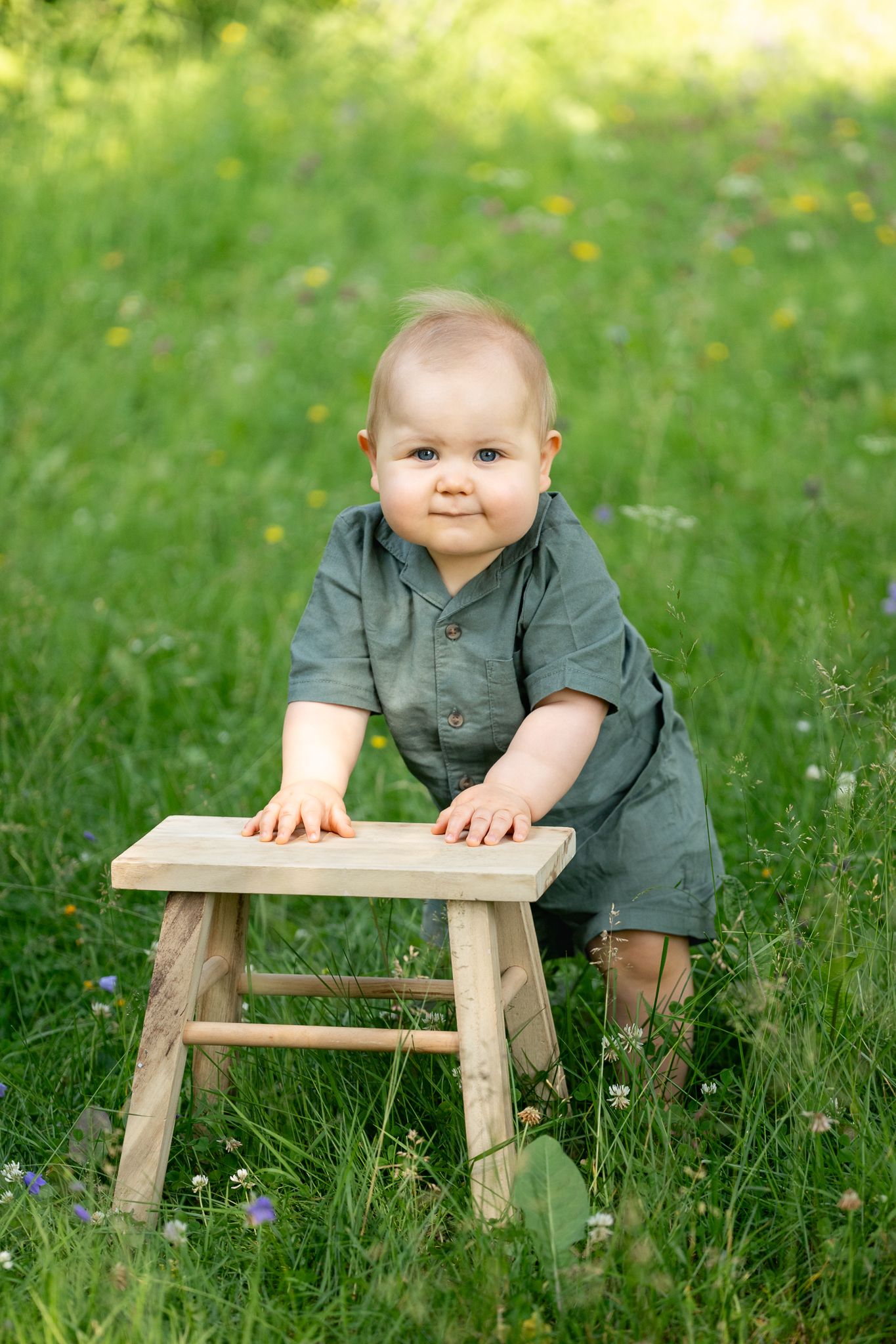 Adorable baby in green romper standing outdoors in a meadow, holding a small wooden stool.