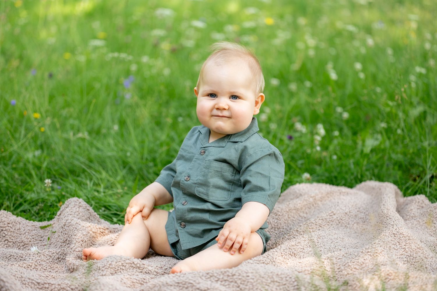 A happy baby sitting on a blanket in green grass, wearing a sage romper, smiling outdoors in summer.