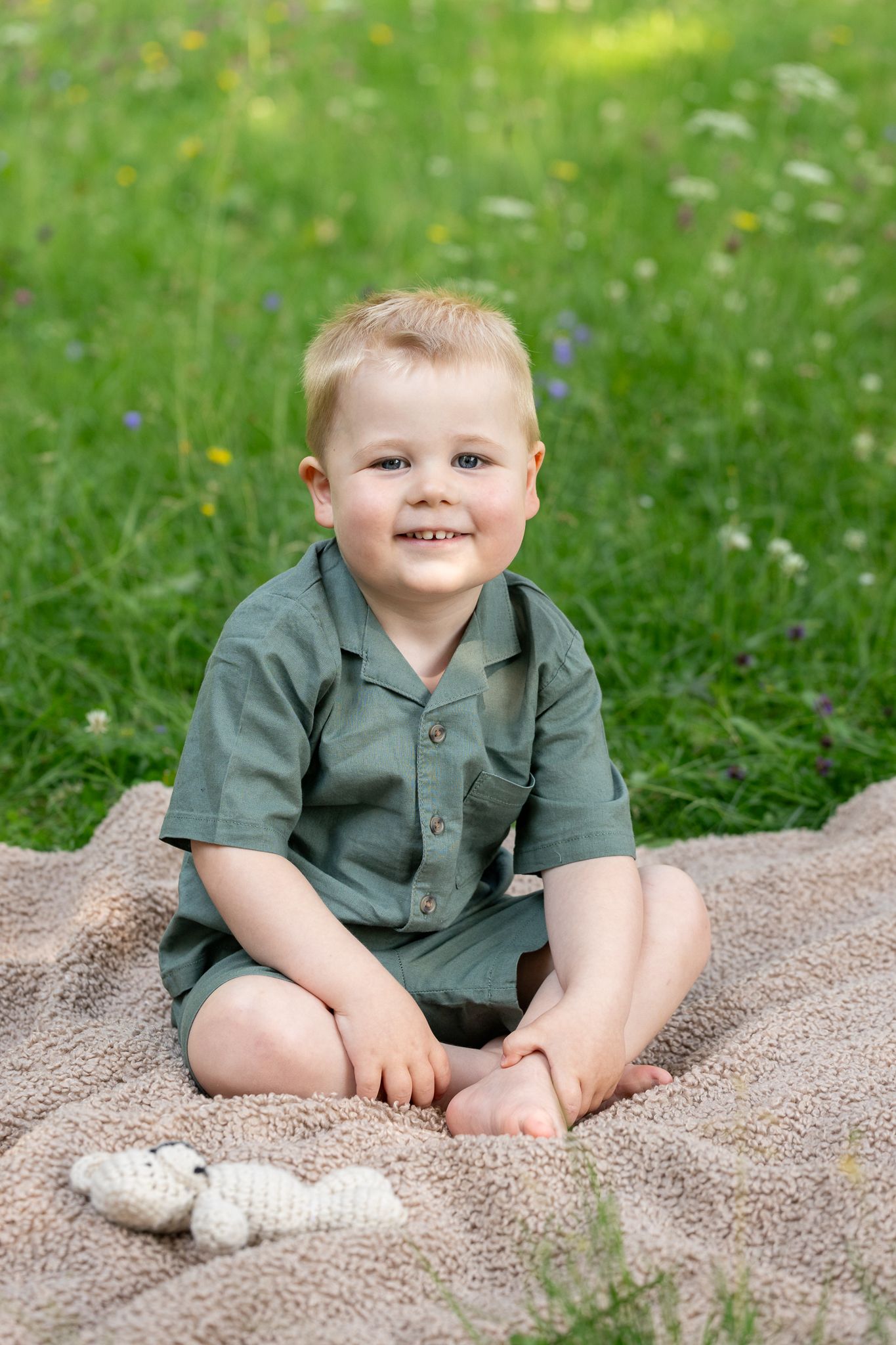 Smiling toddler boy in green outfit sitting cross-legged on blanket in sunny meadow with wildflowers.
