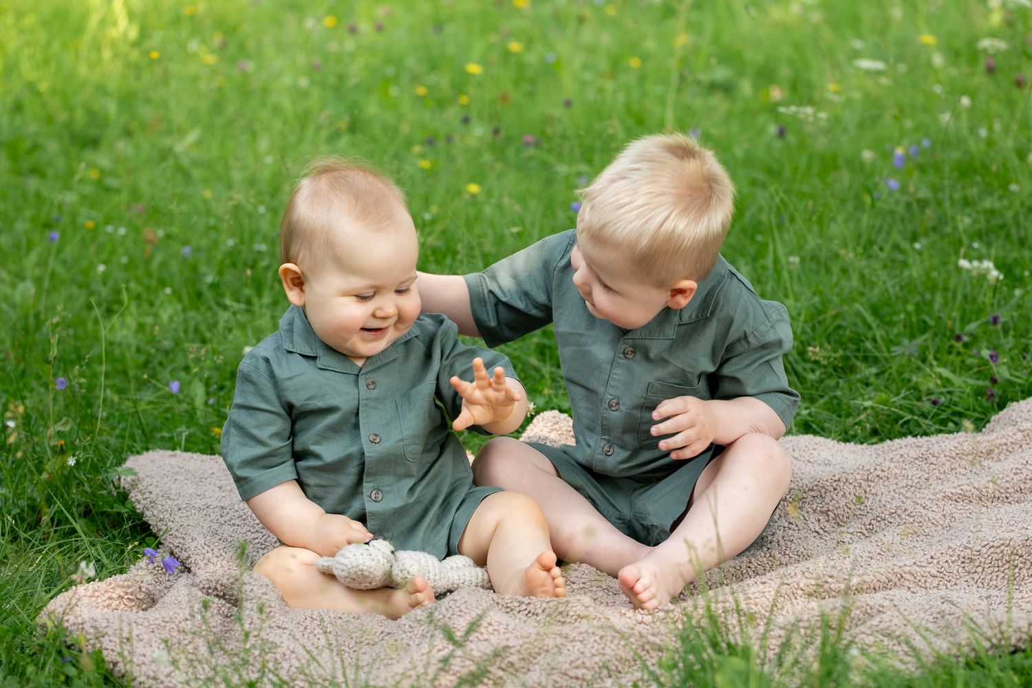 Two baby boys in matching green outfits sitting on a blanket in grass, playing with toys together outdoors.