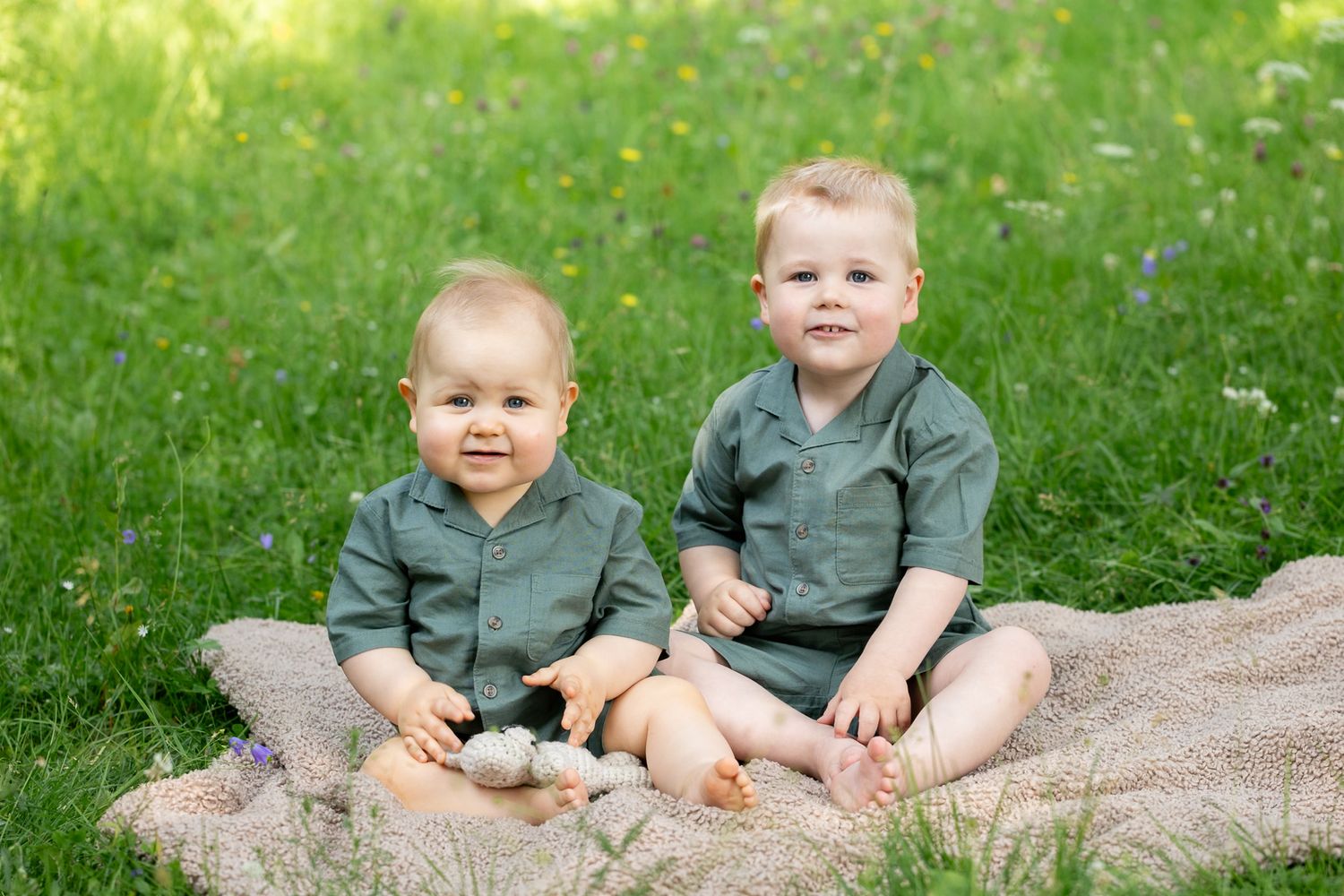 Two adorable toddler boys in matching green outfits sitting on a blanket in a lush green grass field.