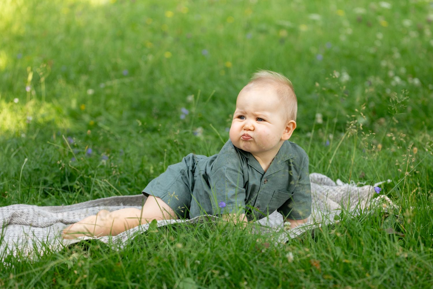 Cute baby in gray outfit making a funny face while lying on a blanket in lush green grass outdoors.