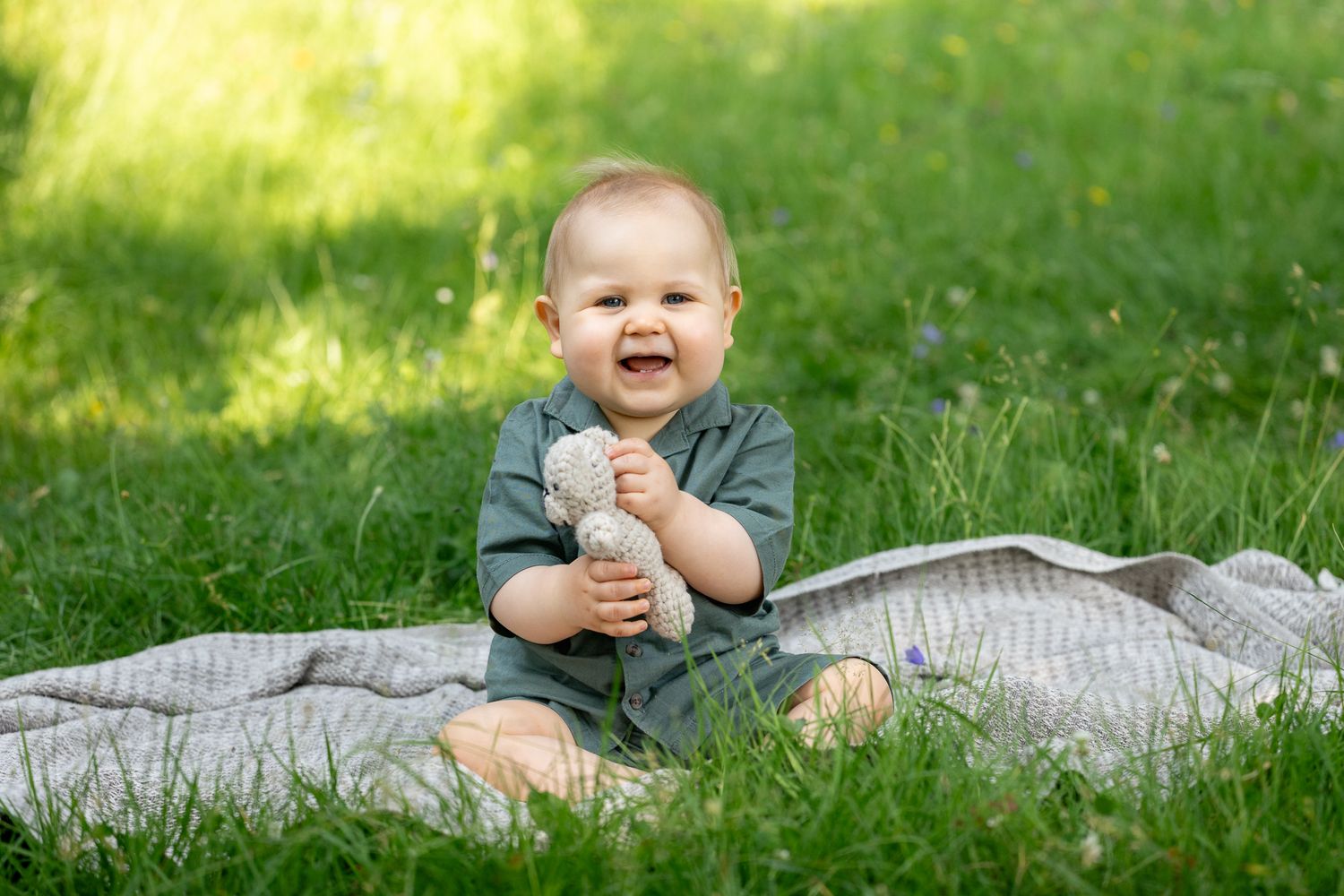 Happy baby boy sitting on a blanket in green grass, smiling and holding a small toy outdoors.