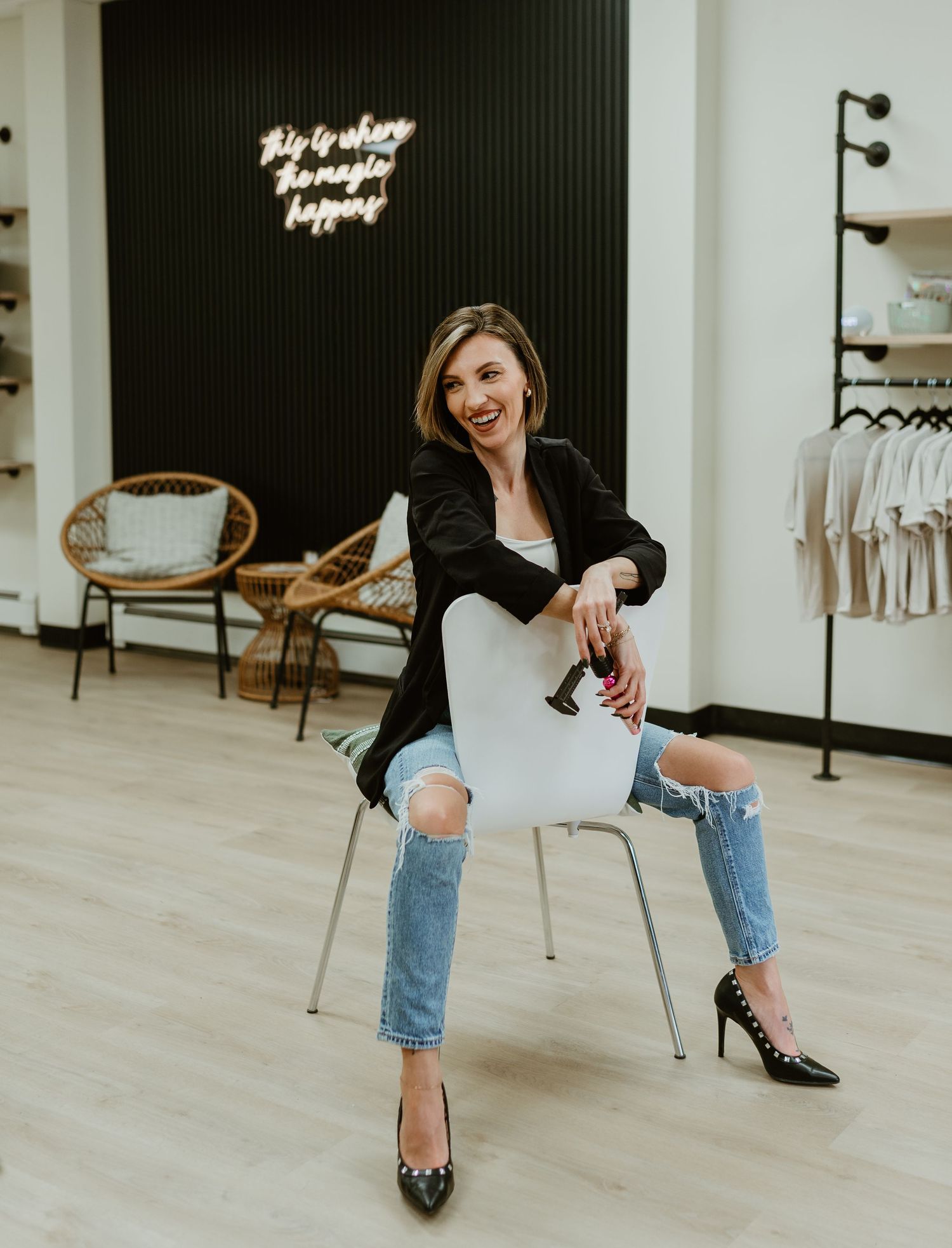 A permanent makeup artist sitting backwards in a chair, laughing confidently in a bright studio, with her tools in hand.