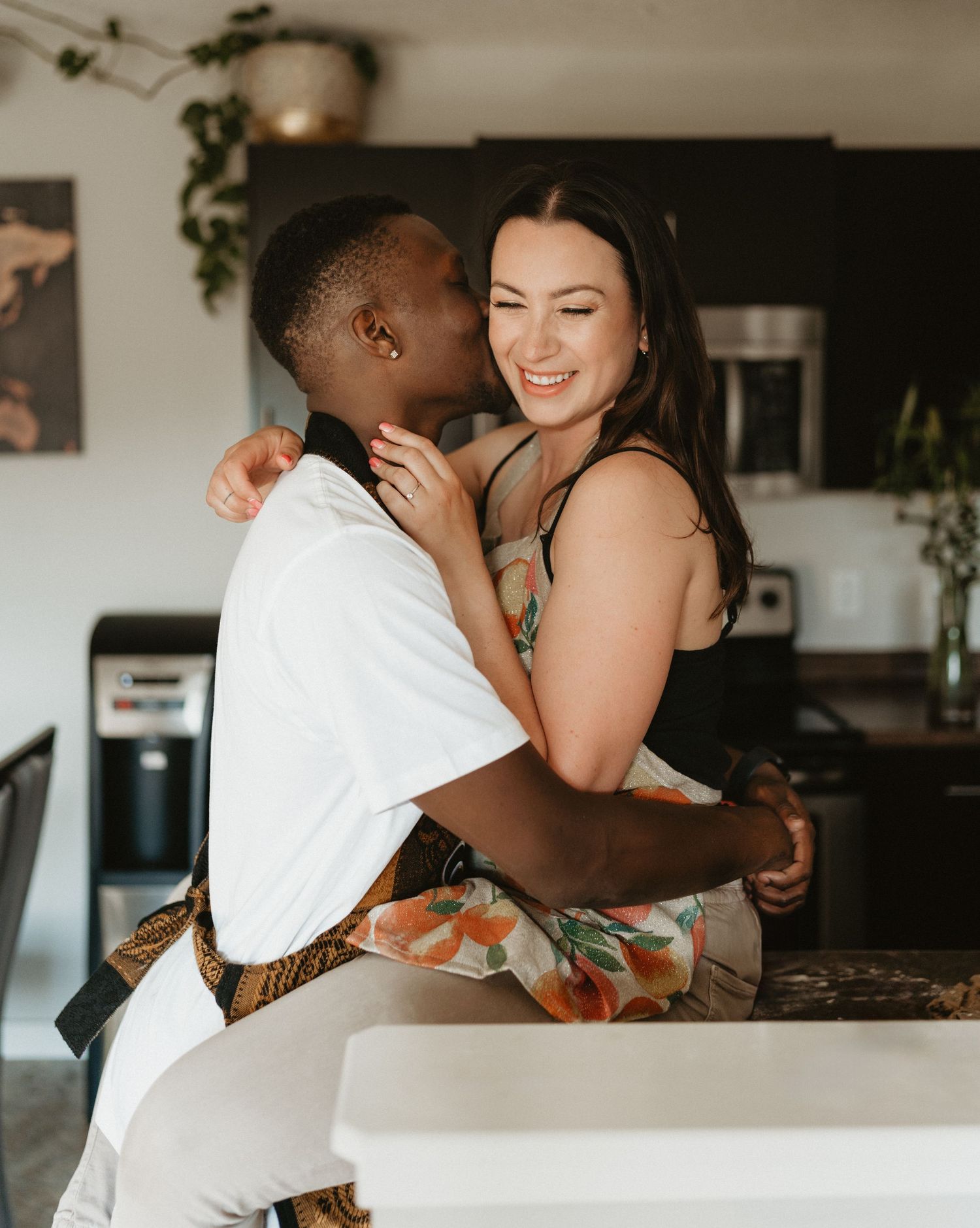 An engaged biracial couple embracing in their kitchen, before making cookies and drinking champagne.