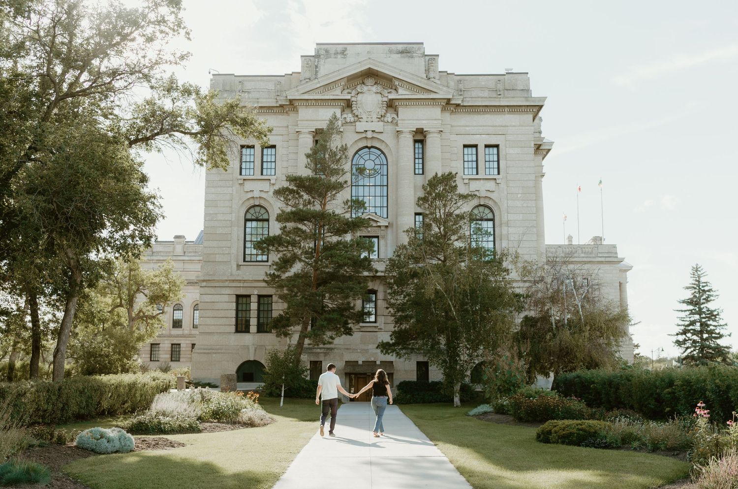 A happy engaged couple walking towards the camera on a sunny day, hand in hand, with a historic building in the background.