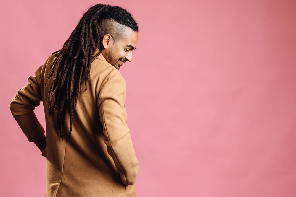 Side profile of person with long dreadlocks wearing tan jacket against pink background.