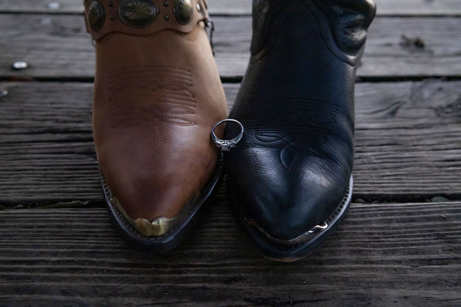 Two cowboy boots, one brown and one black, with a diamond wedding ring balanced between them on a wooden deck.