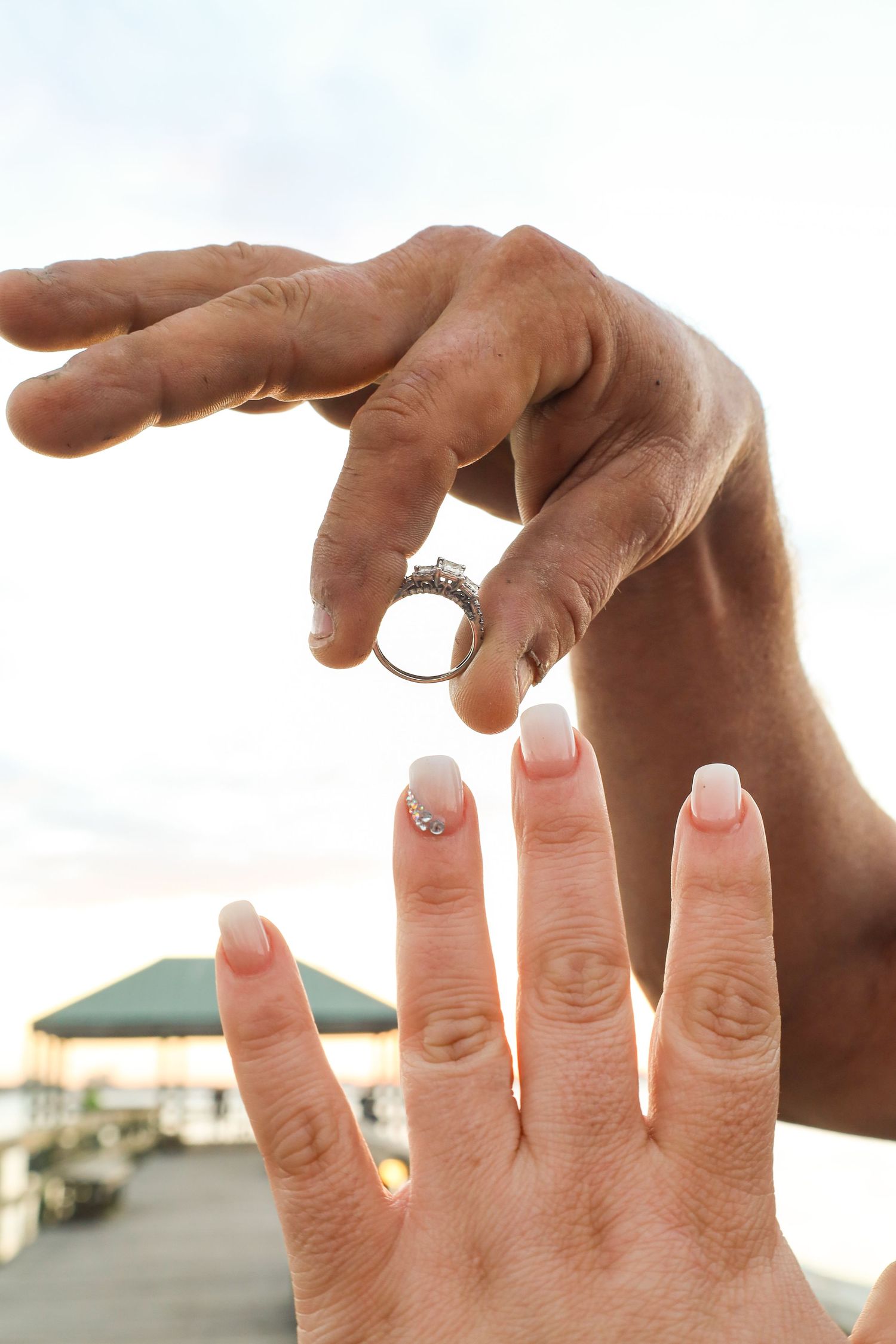 A man holds a diamond engagement ring above a woman's outstretched hand with French manicured nails outdoors.