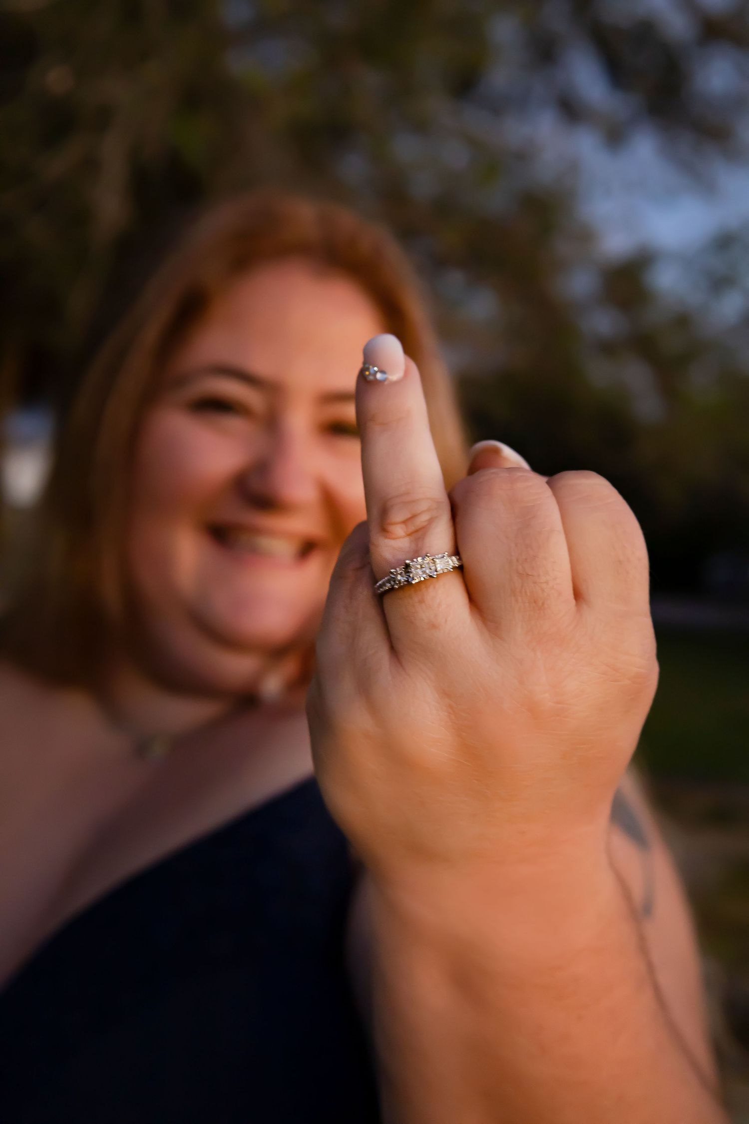 Smiling woman showing off her diamond engagement ring and wedding band outdoors at golden hour.