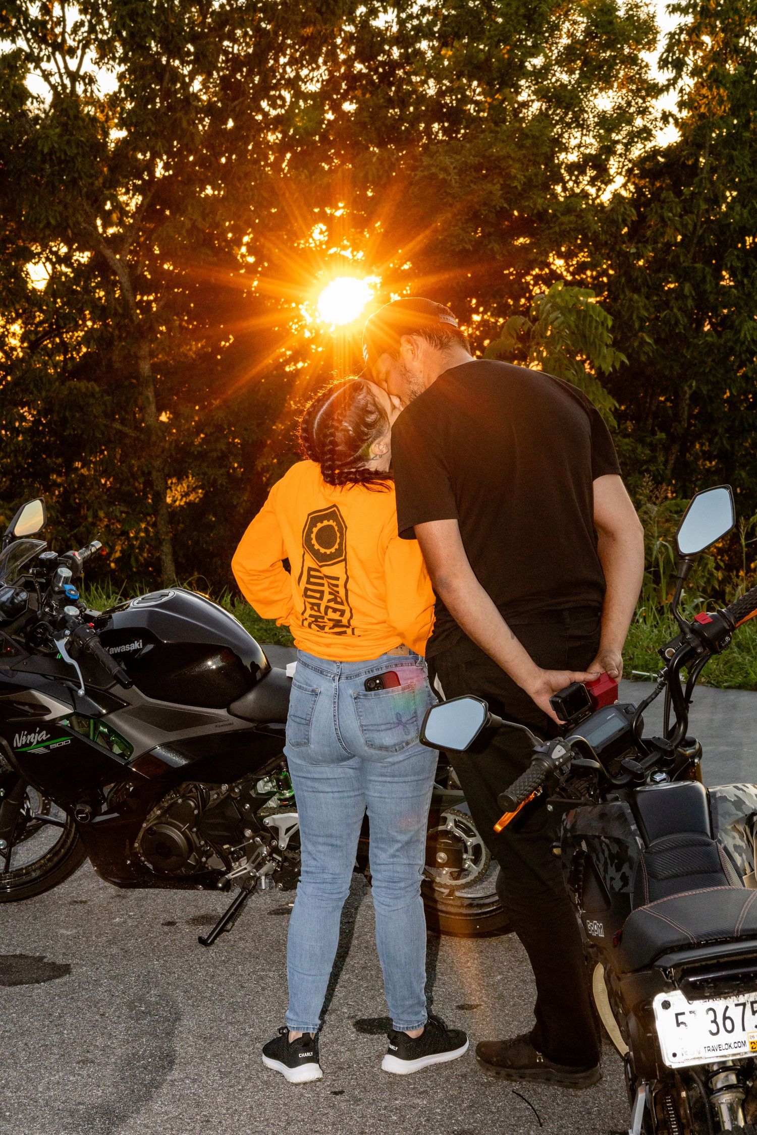 Couple sharing a romantic moment beside motorcycles at golden sunset, woman in orange hoodie and jeans.