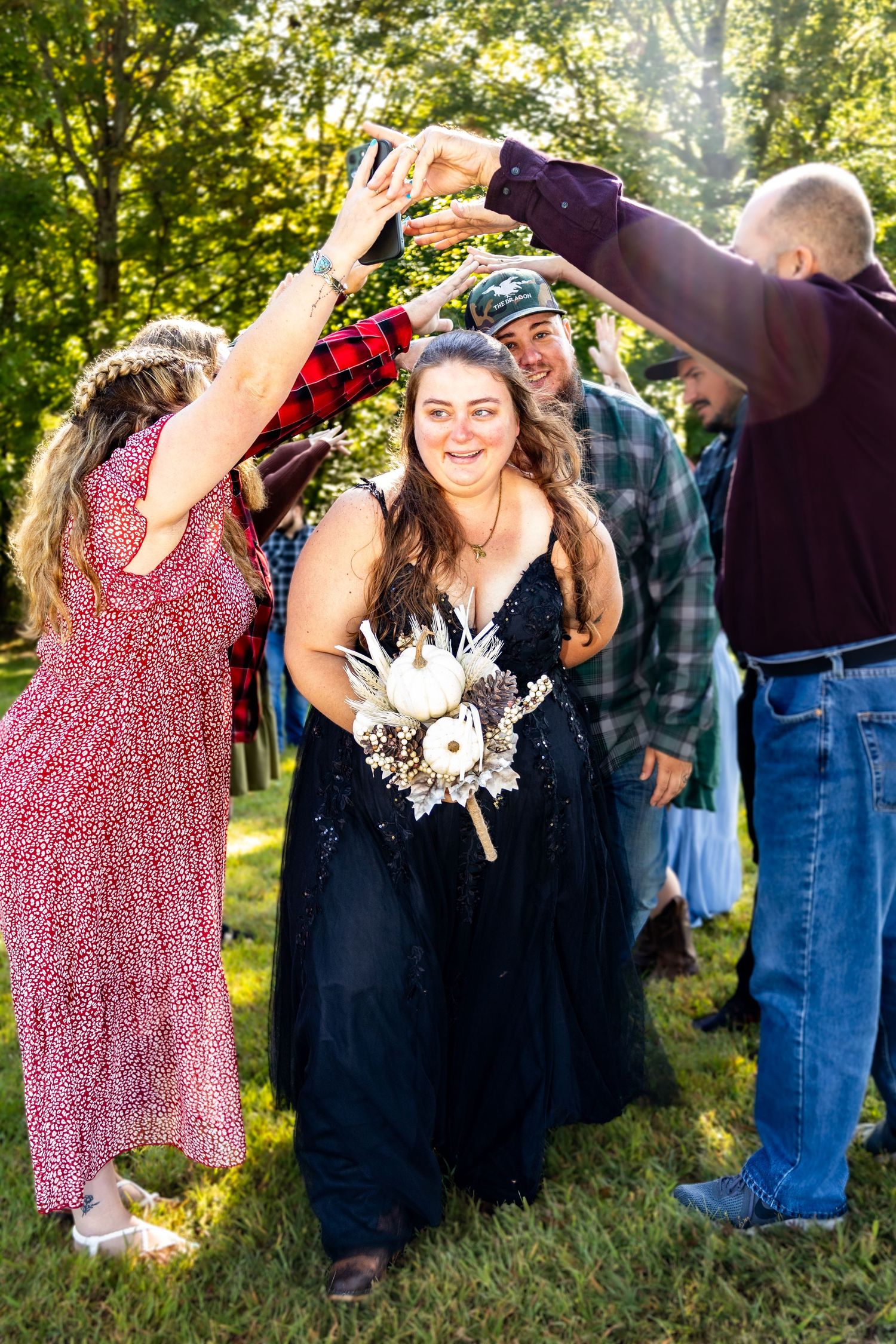 Bride in black sequin gown holds white bouquet while guests form an arch with their hands outdoors.