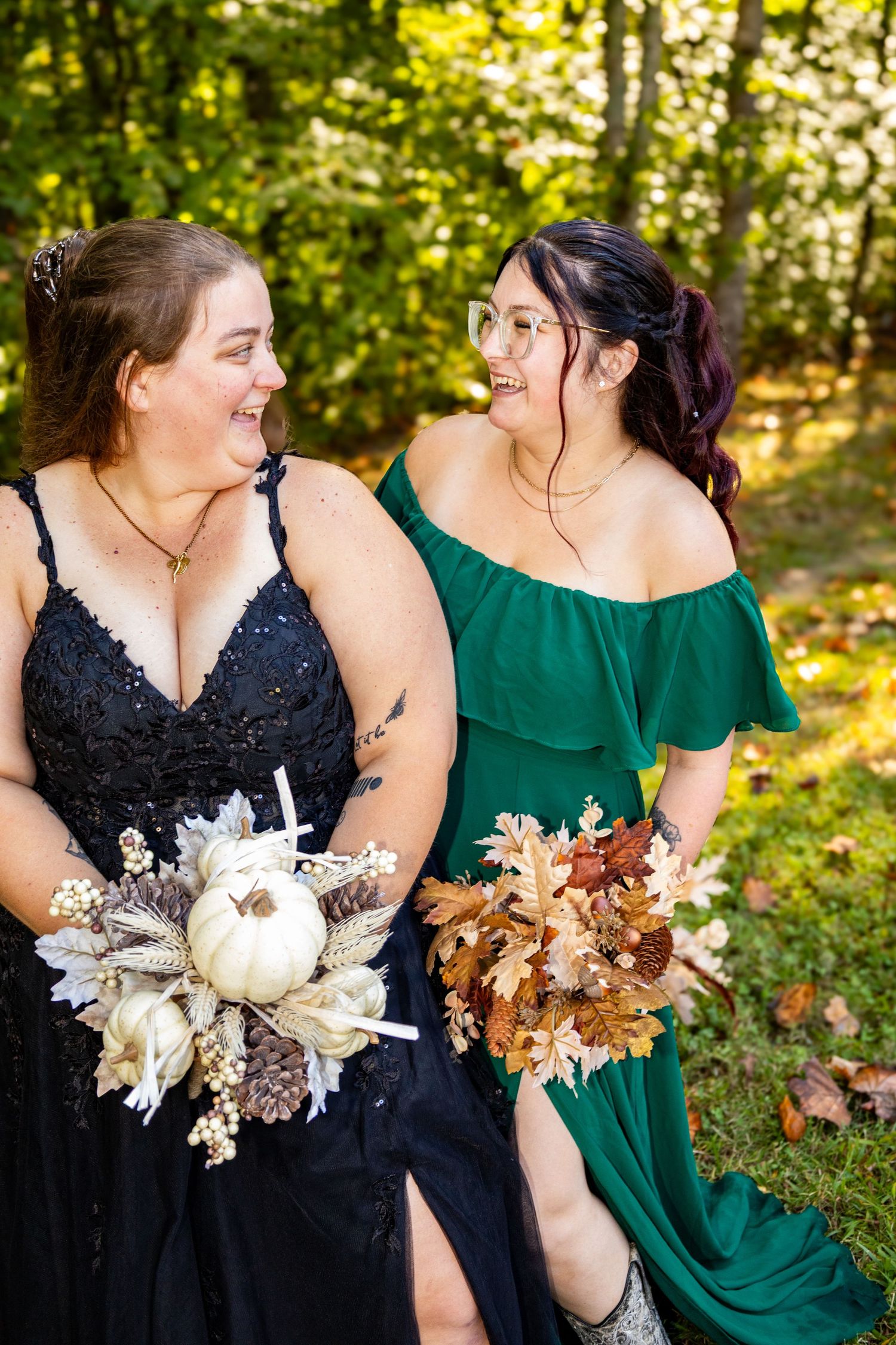 Two bridesmaids in black and green dresses holding autumn bouquets, laughing outdoors at a fall wedding.
