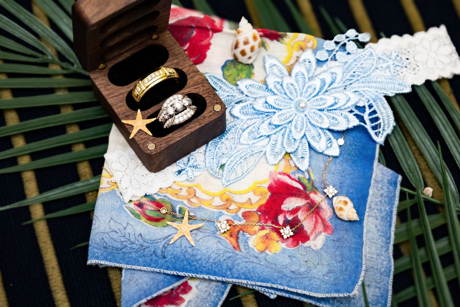Wedding rings in wooden box with blue lace garter and floral handkerchief on tropical palm leaves.