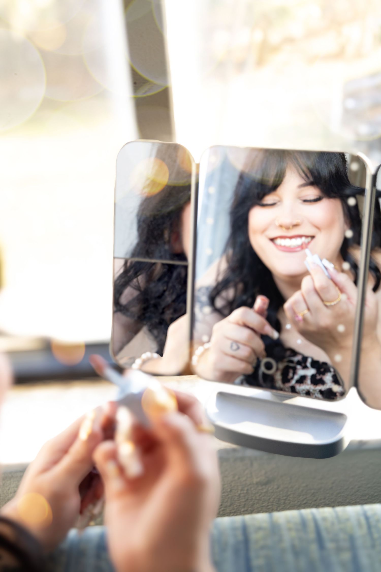 Woman smiling in a trifold vanity mirror while applying lipstick, wearing pearl earrings and a leopard print top.