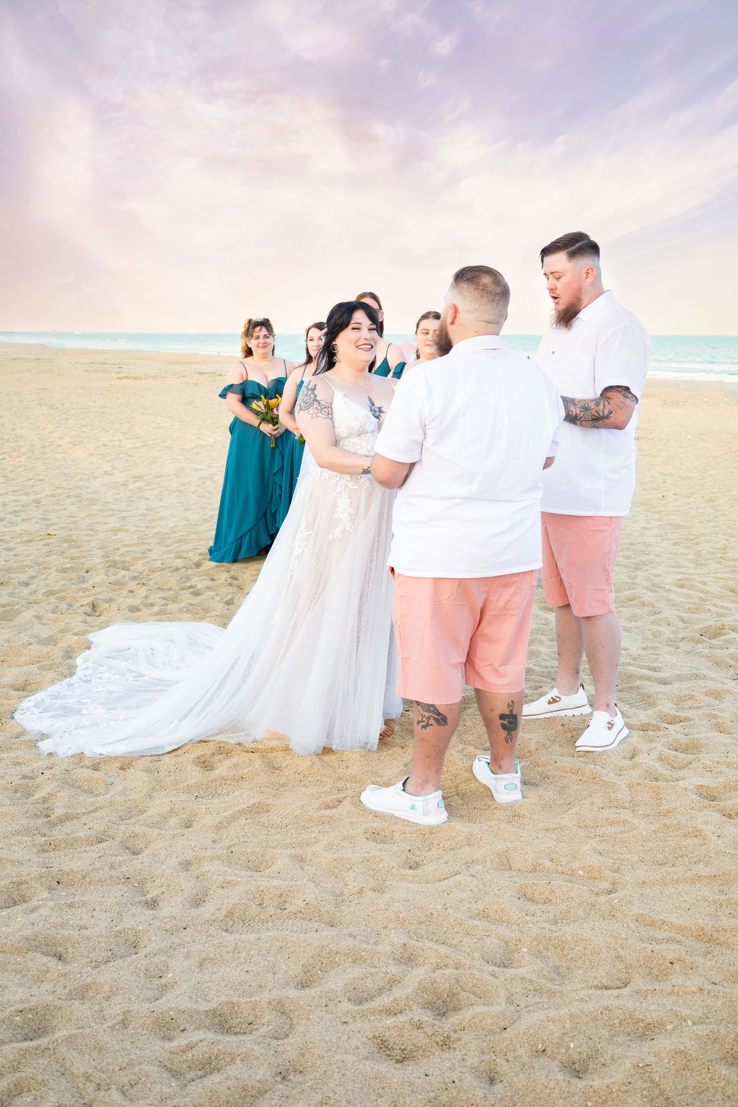 Bride and groom exchange vows on sandy beach at sunset with bridesmaids in teal dresses standing nearby.