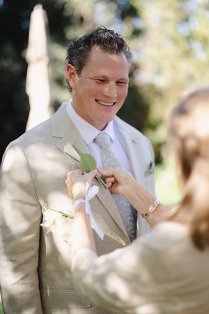 Smiling groom in tan suit receiving boutonniere pin at outdoor wedding ceremony.