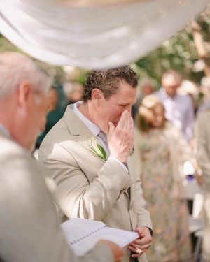 Emotional groom in beige suit covering his face with hands during outdoor wedding ceremony.