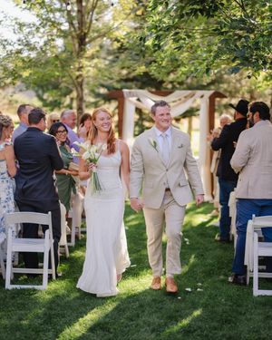 Smiling newlyweds walk down outdoor wedding aisle, bride in white gown holding bouquet, groom in tan suit, guests applauding.