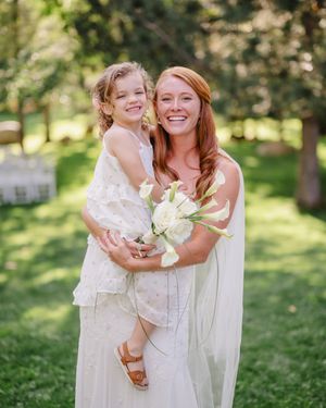 A smiling bride in a white gown holds a young flower girl outdoors, both wearing white at a garden wedding ceremony.