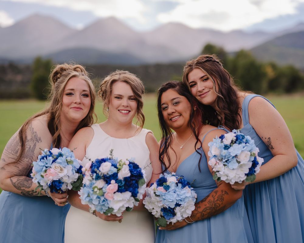 Bride in white gown with three bridesmaids in blue dresses holding blue and white bouquets, mountains in background.