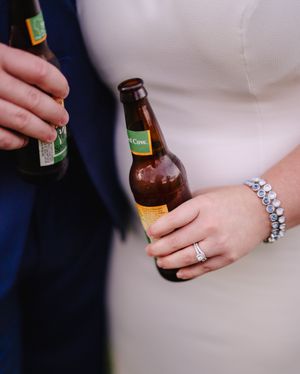 Bride and groom holding beer bottles, showcasing wedding ring and diamond bracelet.