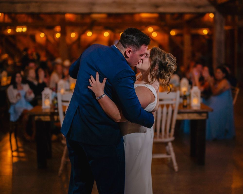 Bride and groom share a romantic first dance kiss in a warmly lit barn wedding reception.