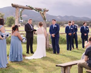 Outdoor wedding ceremony with bride and groom at rustic wooden arch, bridesmaids in blue, groomsmen in navy, mountain backdrop.