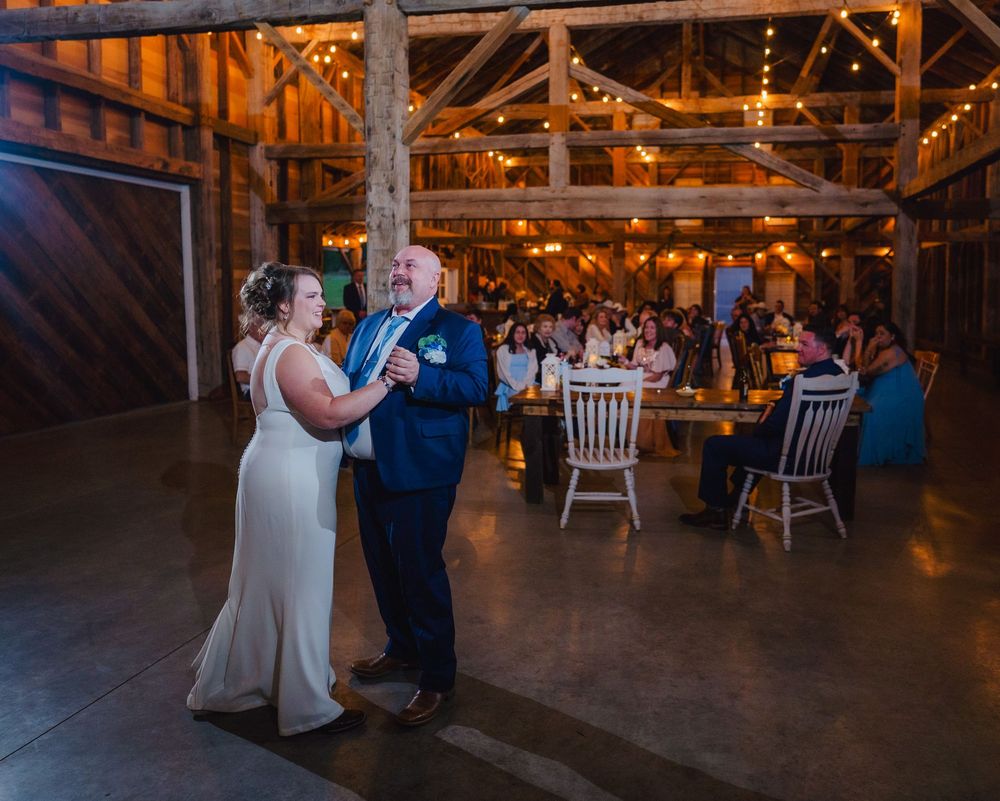Bride and groom share first dance in rustic barn wedding venue with warm string lights and guests seated at tables.