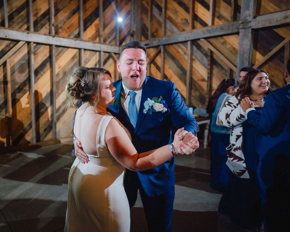 A joyful couple shares an energetic first dance in a rustic barn wedding venue, with guests celebrating in the background.
