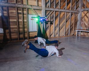 Two people breakdancing on a concrete floor inside a rustic wooden barn with colorful DJ lights overhead.