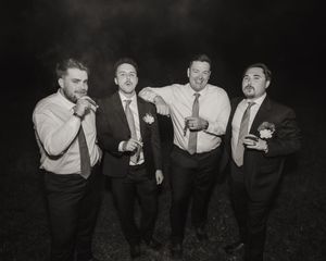 Black and white photo of four groomsmen in suits smoking cigars and laughing together at night.