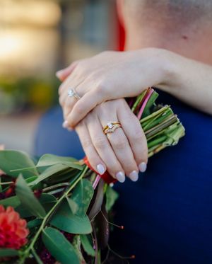 Close-up of a woman's hand with gold engagement ring holding a floral bouquet while embracing a man in a blue suit.