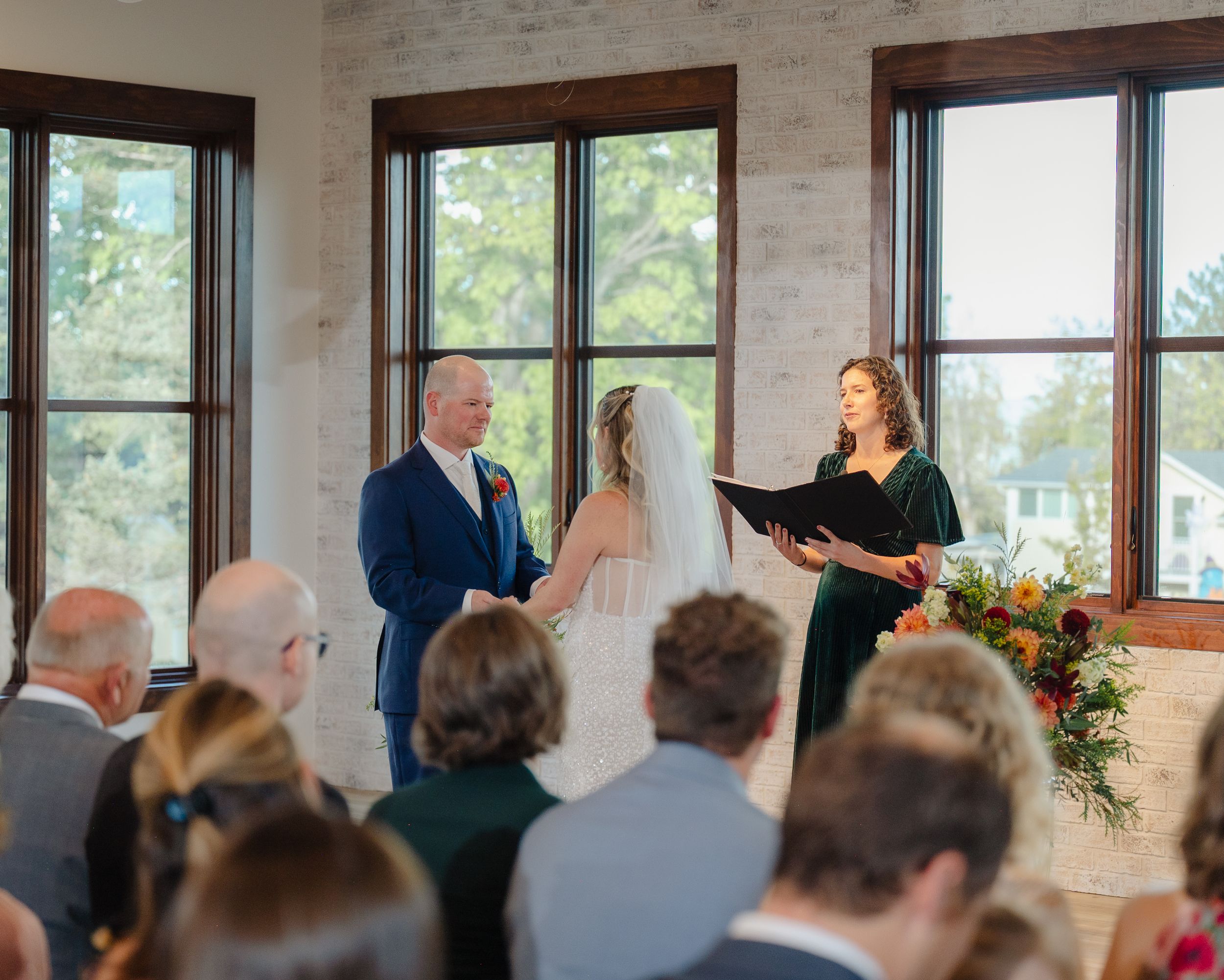 A bride and groom exchange vows at an elegant indoor wedding ceremony with a officiant and guests seated nearby.
