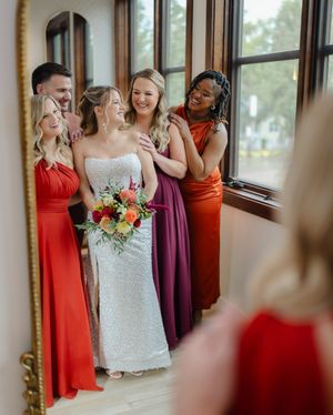 Bride in white sequin gown holds colorful bouquet surrounded by bridesmaids in red, burgundy, and orange dresses before a mirror.