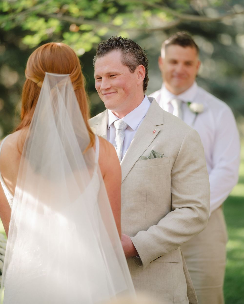 Groom in beige suit smiling at bride during outdoor wedding ceremony with officiant.