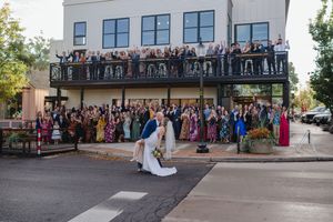 Wedding couple shares romantic dip kiss on street as large crowd of guests cheers from venue balcony and sidewalk behind them.