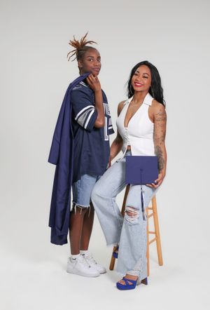 Two stylish women pose together, one in a navy graduation gown and jersey, the other in a white halter top and ripped jeans.
