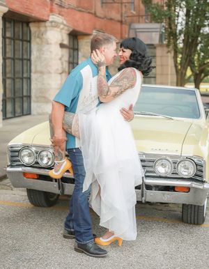A couple in white dress and casual attire share a romantic kiss in front of a vintage yellow car.