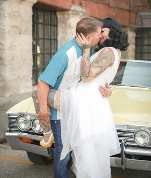 A romantic couple shares a kiss in front of a vintage car, with a white dress and blue shirt creating a retro scene.