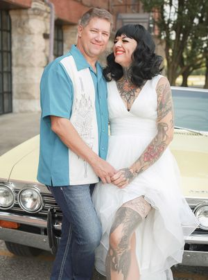 Couple in retro rockabilly style pose together in front of vintage car, bride wearing white dress with visible tattoos.