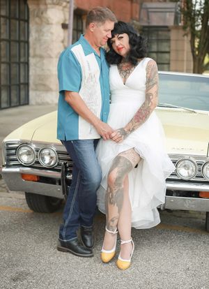 Couple in retro rockabilly style poses by vintage car, bride in white dress shows leg tattoo with yellow heels.